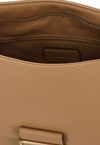 Innenraum einer tanfarbenen Leder-Handtasche mit einem Reißverschlussfach und glatter Textur, feinen Nähten und einem Markenetikett.