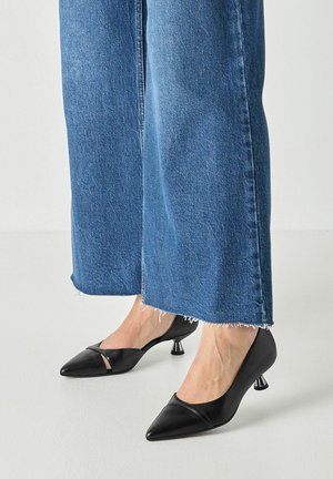 Fødder iført sorte spidse lavhælede sko med metal detalje, kombineret med blå cropped vidde bukser med frynset kant.