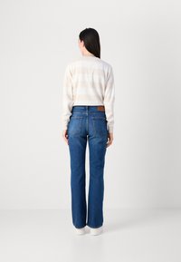 En kvinne står med ryggen til kameraet, iført en stripete genser og blå flared jeans, mot en enkel hvit bakgrunn.