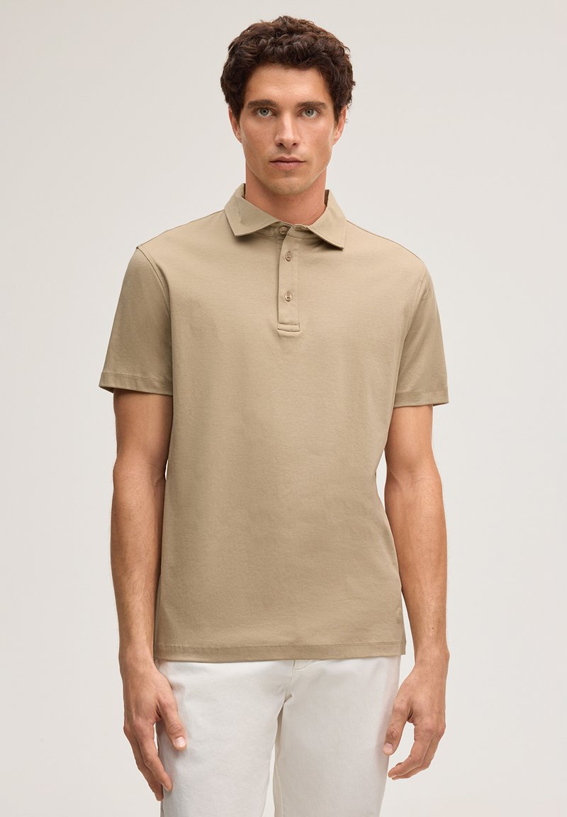 Jonge man met krullend bruin haar, gekleed in een beige poloshirt met korte mouwen en een witte broek, staand tegen een effen lichte achtergrond.