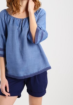 Kobieta w niebieskiej bluzce z odkrytymi ramionami i koronkową lamówką oraz granatowych podwiniętych szortach, stojąca na prostym jasnym tle.