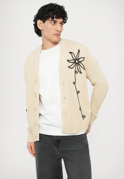 Ελαφρύ μπεζ πλεκτό ζακέτα με σχέδιο λουλουδιού σε μαύρη κεντητική, με έξι κουμπιά και ριμπ μανσέτες. Φορεμένο πάνω από ένα λευκό πουκάμισο.