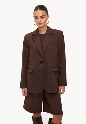 Femme portant un blazer marron oversize et un short assorti longueur genou, accessoirisée de grandes boucles d'oreilles en or et de bagues en or.