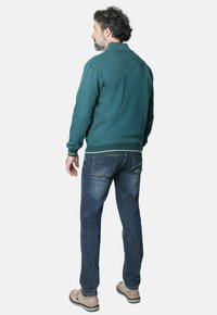 Sudadera verde con puños y dobladillo acanalados, combinada con vaqueros azules. El modelo muestra una vista lateral, con detalles de costura visibles y zapatos casuales.