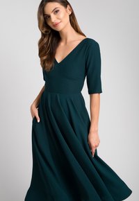Sukienka z dekoltem w serek, krótki rękaw, w głębokim zielonym kolorze. Sylwetka A-line z rozkloszowanym dołem i bocznymi kieszeniami. Gładka faktura, minimalne detale.