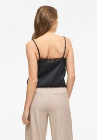 Camisole en satin noir avec bordure en dentelle à l'arrière, bretelles réglables, portée avec un pantalon beige clair sur mesure. Texture lisse, design élégant.