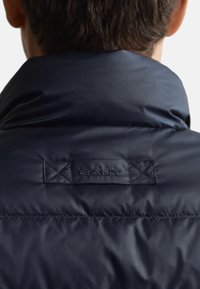 Mörkblå vadderad jacka med hög krage. Har en sydd logotypetikett på baksidan som visar en slät, glänsande textur.