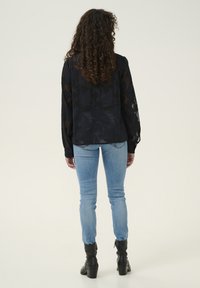 Blusa nera trasparente con motivo floreale, maniche lunghe, polsini a bottoni; abbinata a jeans skinny blu chiaro e stivaletti neri con tacco.