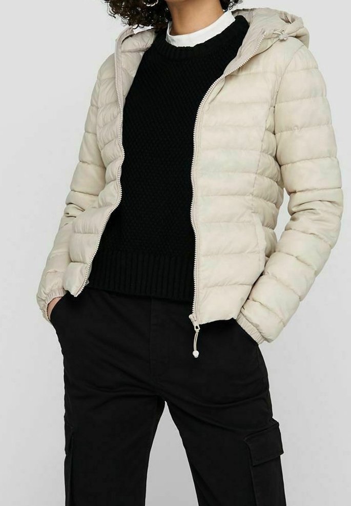 Doudoune beige avec capuche, fermeture éclair et design matelassé. Portée sur un pull noir en tricot et associée à un pantalon cargo noir.