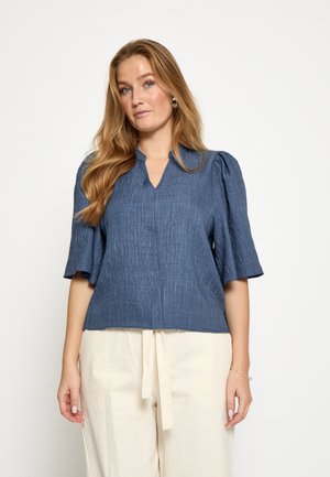 Blouse bleu foncé à manches courtes avec un col en V et un tissu texturé. Caractérisée par des manches légèrement bouffantes et une coupe décontractée. Portée avec un pantalon de couleur claire.