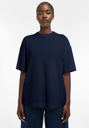 Jeune femme noire debout, portant une chemise ample à manches courtes bleu marine foncé et un jean bleu foncé sur un fond clair uni.