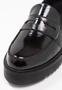 Mocassin en cuir verni noir avec un bout arrondi, présentant des détails cousus et une semelle épaisse en caoutchouc noir. Texture lisse et brillante.
