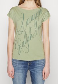T-shirt en coton vert avec un large col et des manches courtes, ornée d'un texte en écriture cursive dans un vert plus foncé. Associé à un jean en denim bleu.