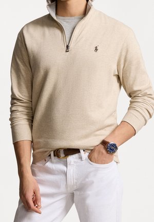 Mann trägt beige Viertel-zip Pullover mit Polo-Logo, weiße Hose, braunen Gürtel und blaues Armband mit braunem Lederarmband, Hand in der Tasche.