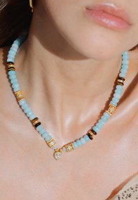 Collier perlé avec des perles bleu clair, des accents dorés et des pièces circulaires sombres. Un pendentif en cristal en forme de goutte est mis en valeur.