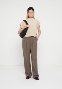 Beige short-sleeve top and brown high-waisted trousers, standing with a black shoulder bag. Textured fabric with a relaxed fit.