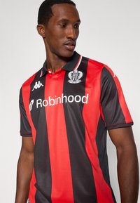 Μπλούζα ποδοσφαίρου με μαύρες και κόκκινες ρίγες, με γιακά πόλο, λογότυπο Kappa και έμβλημα συλλόγου στο στήθος. Το υλικό φαίνεται λείο και διαπνέον.