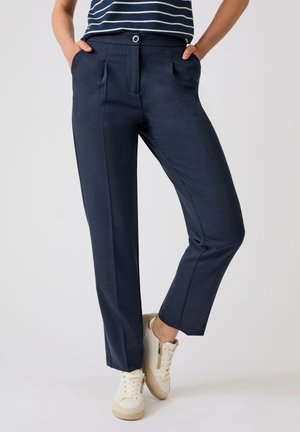 Persoon draagt marineblauwe op maat gemaakte broek, witte sneakers met beige zolen en een marineblauw-wit gestreept overhemd, staand met handen in de zakken.