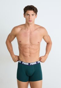 Boxers verdes de algodão com uma cinta azul-marinho apresentando a marca "PUMA" em branco. O modelo tem um corpo atlético e está sem camisa.