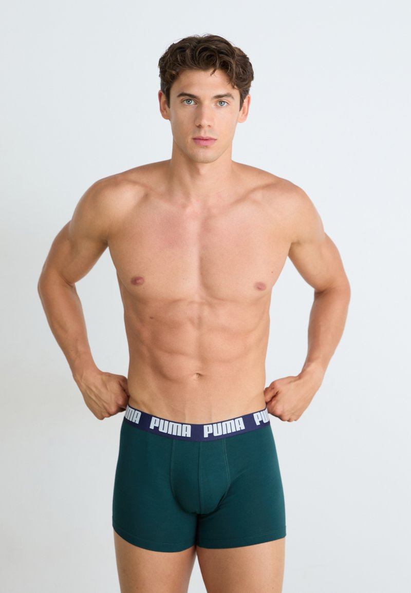 Boxers verdes de algodão com uma cinta azul-marinho apresentando a marca "PUMA" em branco. O modelo tem um corpo atlético e está sem camisa.