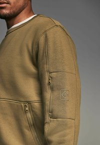 Sudadera verde oliva con un acabado texturizado, que cuenta con un bolsillo lateral con cremallera y un parche en la manga con un logo de diamante.