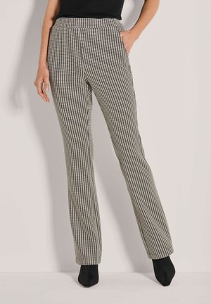 Vrouw in hoge taille, rechte pijpen met een houndstooth-patroon, met één hand in de zak en zwarte hoge laarzen.