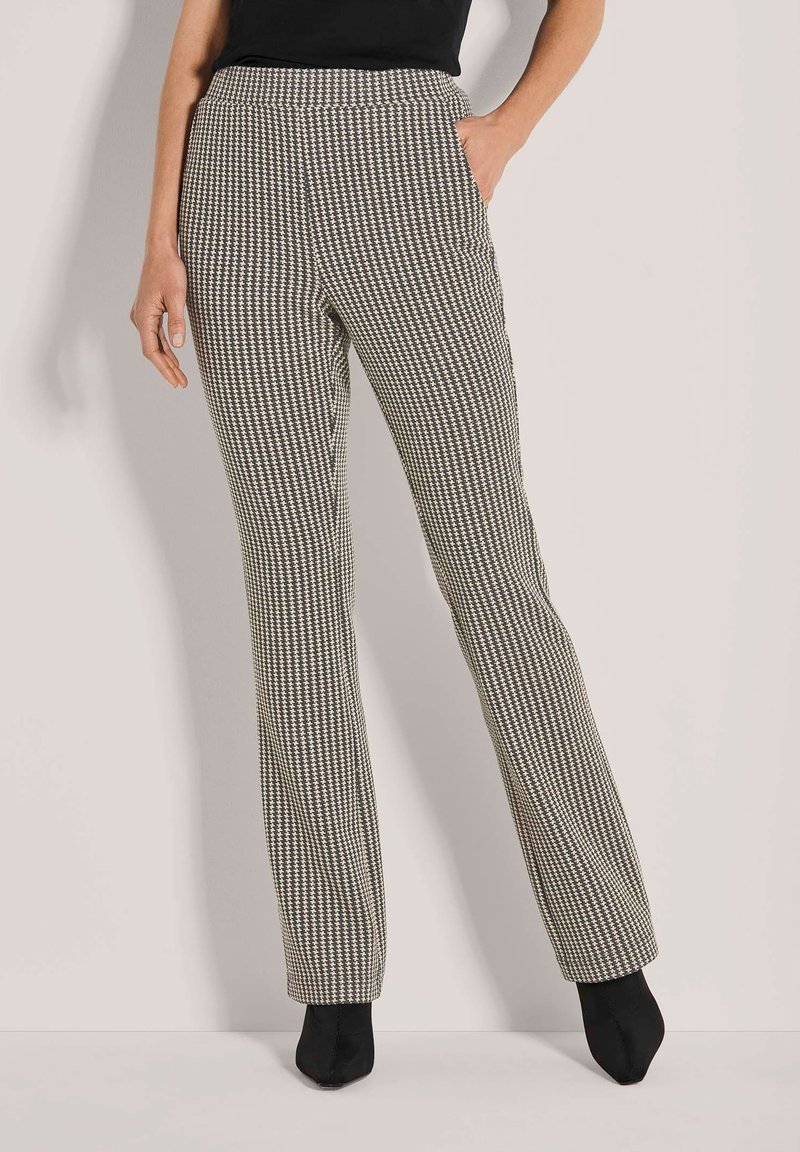 Vrouw in hoge taille, rechte pijpen met een houndstooth-patroon, met één hand in de zak en zwarte hoge laarzen.