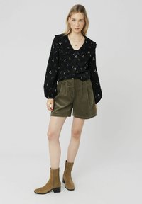 Chemise noire avec un motif floral et un col froncé, associée à un short en velours côtelé vert olive. Des bottines marron complètent la tenue.