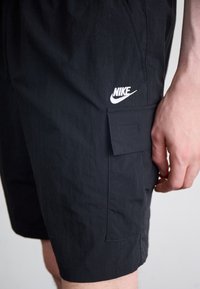 Čierne cargo šortky Nike s textúrovanou tkaninou, bočnou kapsou a bielym logom. Dizajn má uvoľnený strih a viditeľné švy.