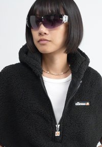 Veste zippée en polaire noire avec un col haut et un petit logo. Le modèle porte des lunettes de soleil teintées de violet avec des accents en forme d'étoile et un t-shirt blanc.