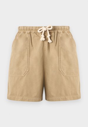 Ung mann med krøllete hår iført en lys gråstripet oversized T-skjorte, beige shorts med snor, hvite sokker og hvite joggesko, stående stille.