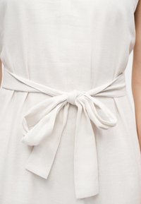 Λευκή υφασμάτινη ζώνη δεμένη σε φιόγκο στη μέση ενός ανοιχτόχρωμου φορέματος, που δείχνει την υφή και τις απαλές πτυχώσεις του υφάσματος.