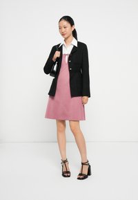 Zwarte textuur blazer over een roze A-lijn jurk met een witte kraag; gecombineerd met zwarte sandalen met banden. Minimaal accessoiregebruik, eenvoudig ontwerp.