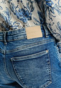 Zbliżenie na tył niebieskich dżinsów z beżową metką "greenpoint" w zestawieniu z białą bluzką w niebieski kwiatowy wzór.