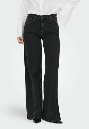 Femme portant un jean noir taille haute à jambes larges avec poches avant, associé à un haut blanc à manches longues et des chaussures noires pointues.