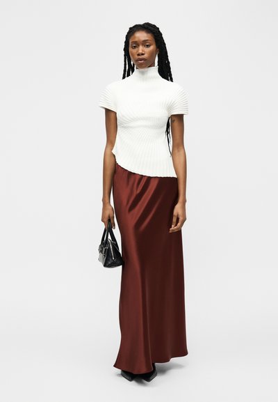 Femme aux longs cheveux nattés portant un haut à col roulé blanc côtelé à manches courtes et une jupe longue en satin marron, tenant un petit sac à main noir.