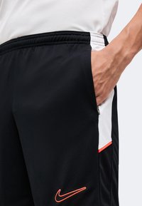 Άτομο που φοράει μαύρα σορτς Nike με πορτοκαλί λογότυπο και λευκές πλευρικές επιφάνειες, με το χέρι μέσα στην δεξιά τσέπη.