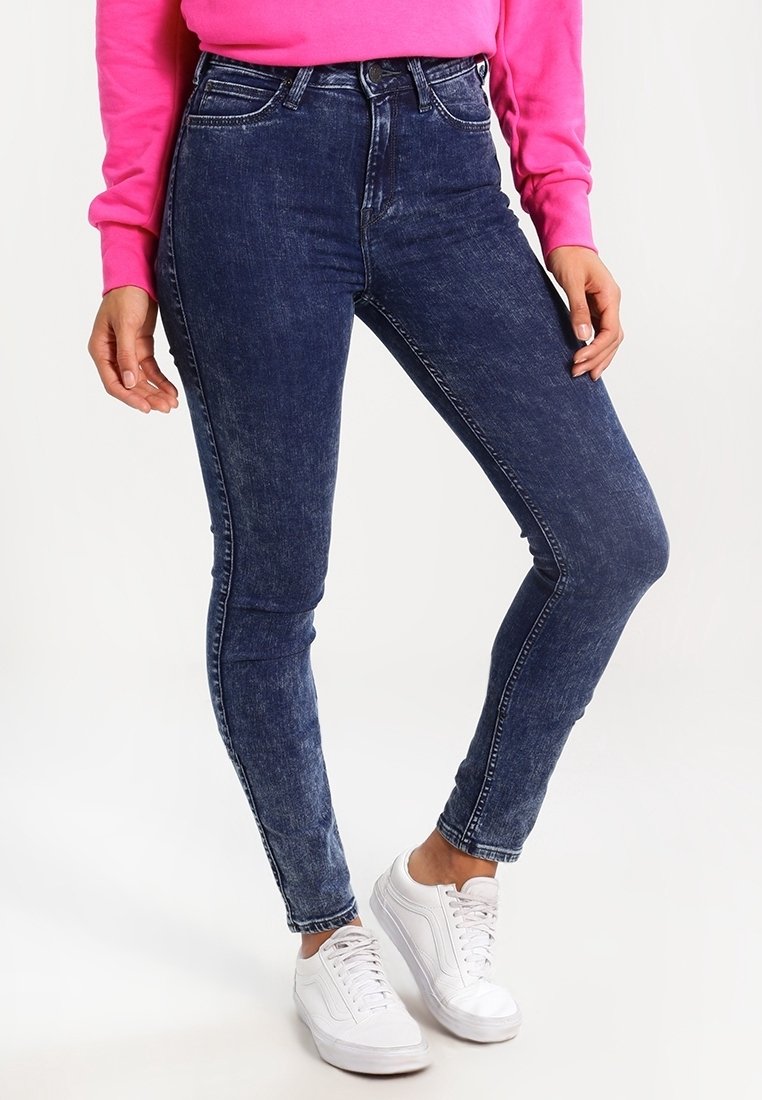 Vaqueros ajustados de medio tiro en un azul oscuro con un acabado descolorido, que presentan un estilo clásico de cinco bolsillos y un corte ajustado. Combinados con zapatillas blancas.