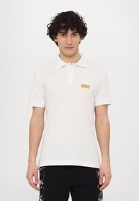 Bela polo majica s ovratnikom, kratkimi rokavi in zlatim logotipom na levi strani prsnega koša. Gladka tekstura in prilegajoča oblikovanje.