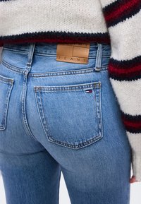 Κοντινή λήψη μπλε τζιν τζιν με πίσω τσέπες και καφέ δερμάτινο έμπλαστρο, φορεμένα με λευκό πουλόβερ που έχει κόκκινες και μαύρες ρίγες στα μανίκια.