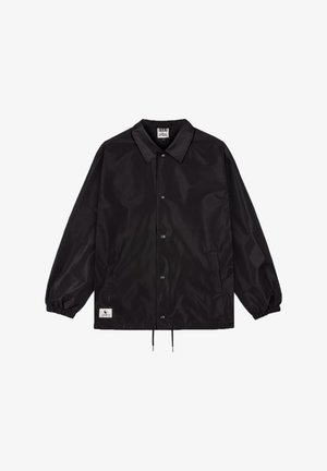 Zwarte nylon jas met lange mouwen, een kraag, drukknopen aan de voorkant, elastische manchetten en een trekkoord onderaan; heeft een merketiket aan de onderkant.