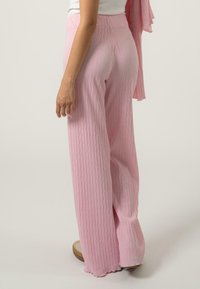 Pantalons larges à côtes rose clair avec une taille haute, présentant une surface texturée et une coupe décontractée, associés à un haut blanc et des sandales.