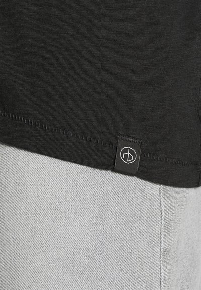 Must musta tekstuuriga must T-särk, mille puusal on logo silt. Allpool särki on nähtav hall denimpakend.