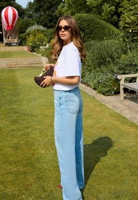 Model trägt ein weißes T-Shirt, hellblaue weite Jeans, hält eine braune geflochtene Tasche, mit Sonnenbrille und langen, welligen Haaren. Grüner Garten im Hintergrund.