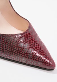Gros plan d’une chaussure à talon haut à bout pointu avec une texture brillante façon cuir de serpent bordeaux et une doublure intérieure beige.