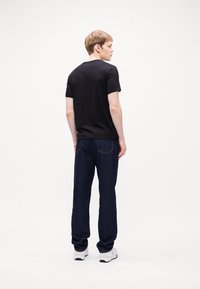 Black short-sleeve t-shirt paired with dark denim jeans. The model wears white sneakers and faces away from the camera, showing a casual look.