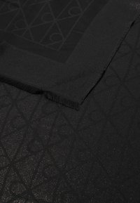 Fekete textil sima felülettel, diszkrét geometriai mintával és rojtos szélekkel. A design különböző textúrájú szakaszokat tartalmaz.