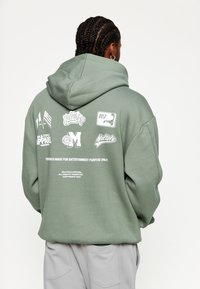 Olivgrüner Hoodie mit großer Kapuze, versehen mit verschiedenen weißen Grafikdrucken und Text auf dem Rücken. Hergestellt aus weichem, strukturiertem Stoff.
