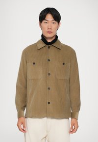 Muškarac u bež korduroj košulji sa dugim rukavima preko crnog rolka i svijetlim krem hlačama, stoji uz jednostavnu bijelu pozadinu.