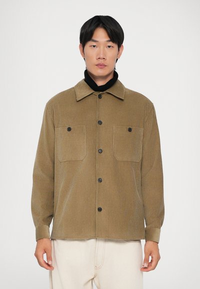 Uomo che indossa una camicia button-up in corda color sabbia sopra un dolcevita nero e pantaloni di un leggero color crema, che si trova in piedi davanti a uno sfondo bianco semplice.